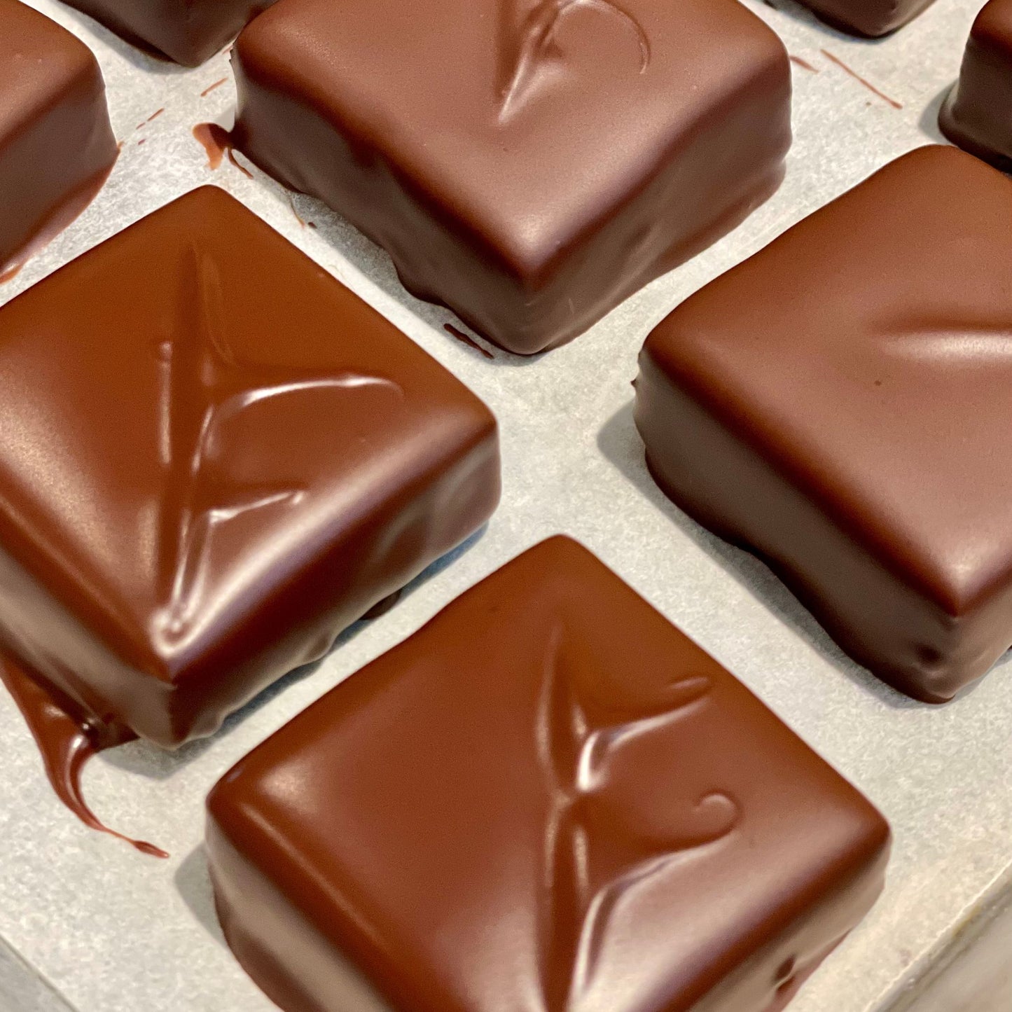 dark chocolate square bonbons, with decorative swirl, some still setting with wet appearing chocolate on parchment paper.