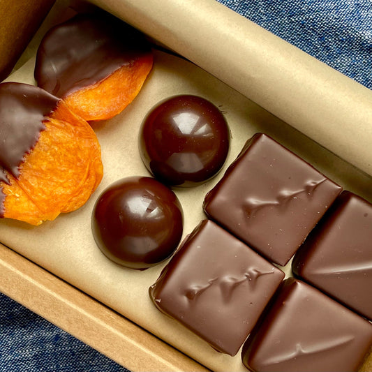 Overhead view of dipped apricots, chocolate domes, dark chocolate square bonbons inside kraft gift box on a denim blue material background.