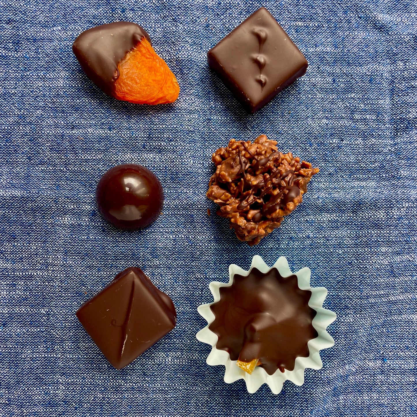 Overhead view of 6 items on denim blue material background: chocolate dipped apricot, chocolate dome, rocher, hazelnut + candied orange cup, 2 dark chocolate bonbons.