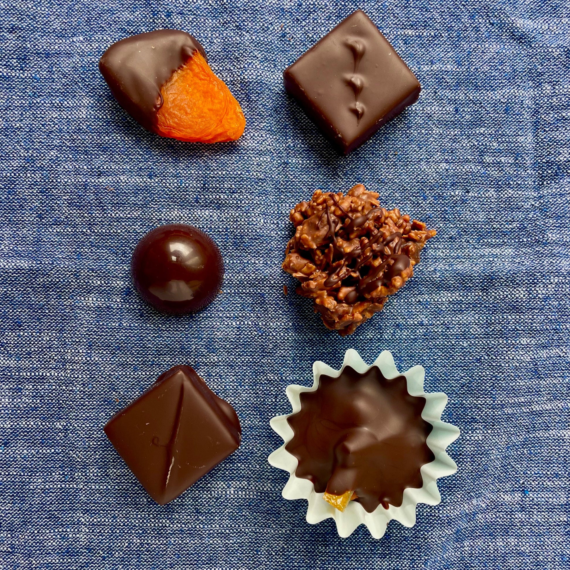 Overhead view of 6 items on denim blue material background: chocolate dipped apricot, chocolate dome, rocher, hazelnut + candied orange cup, 2 dark chocolate bonbons.