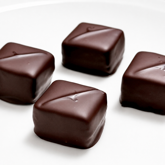 Four square dark chocolate truffles with a glossy finish, arranged on a plain white surface.