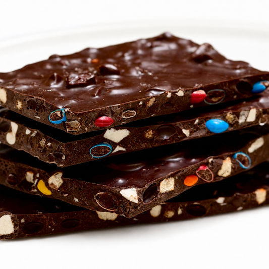 A close-up stack of chocolate bark pieces with  pretzel pieces, and colorful candy-coated bits on a white background.