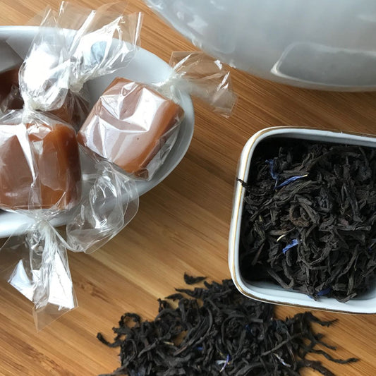 wrapped caramels, loose black tea with blue flowers, sitting in separate white containers on top of a bamboo cutting board background