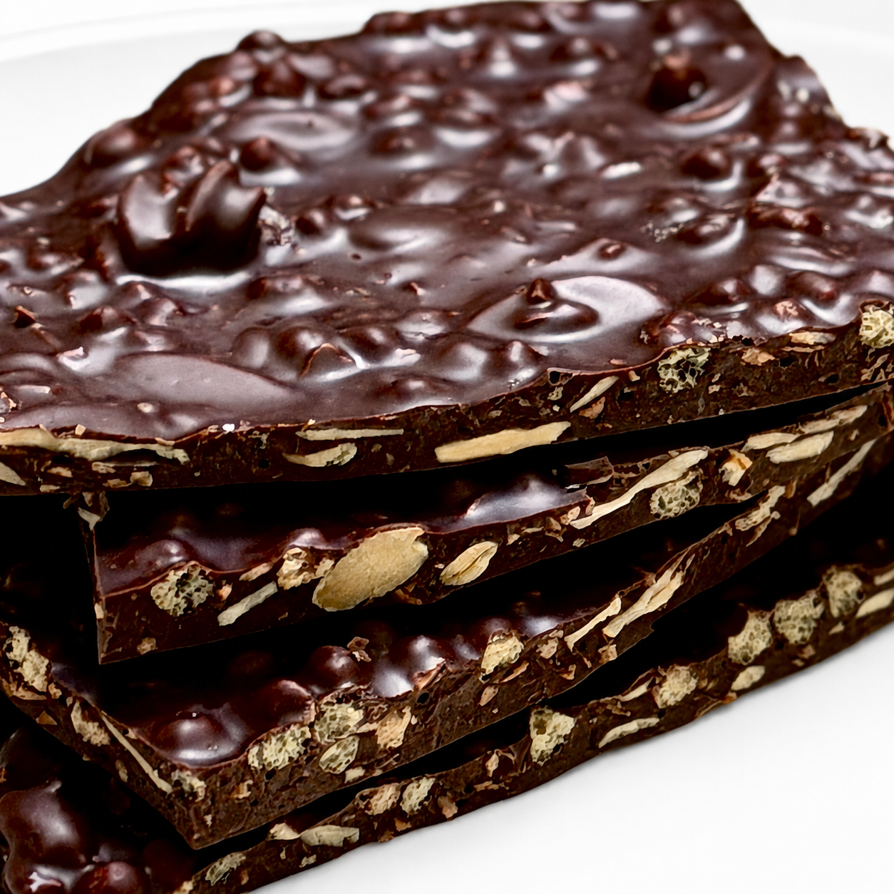 A close-up stack of thick chocolate bark bars with visible nuts and puffed quinoa pieces embedded in glossy dark chocolate, photographed on a white surface.