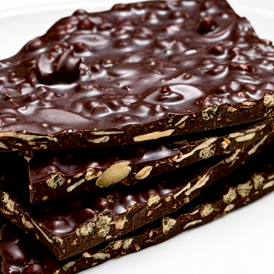 A close-up stack of thick chocolate bark bars with visible nuts and puffed quinoa pieces embedded in glossy dark chocolate, photographed on a white surface.