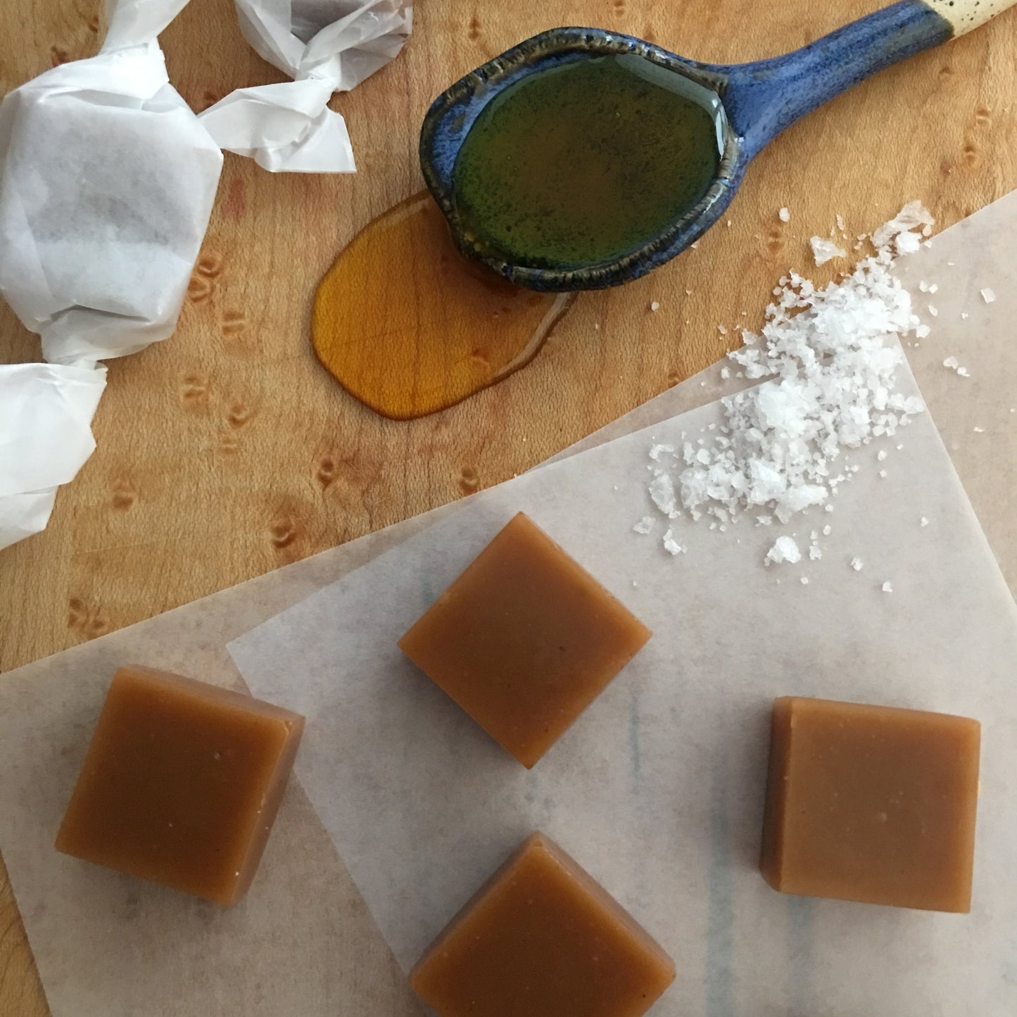 Caramel squares on a wood surface with a ceramic spoon filled with honey spilling out, wrapped caramels,  and white paper in the background.