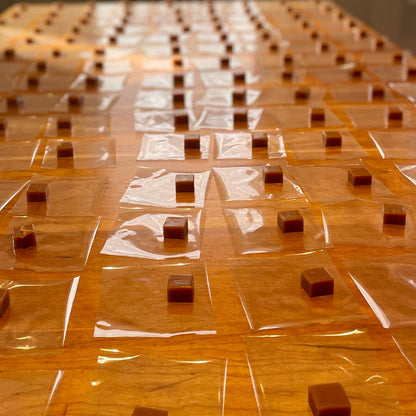 Small square caramels on clear cellophane square sheets on a wooden table, all in a row.