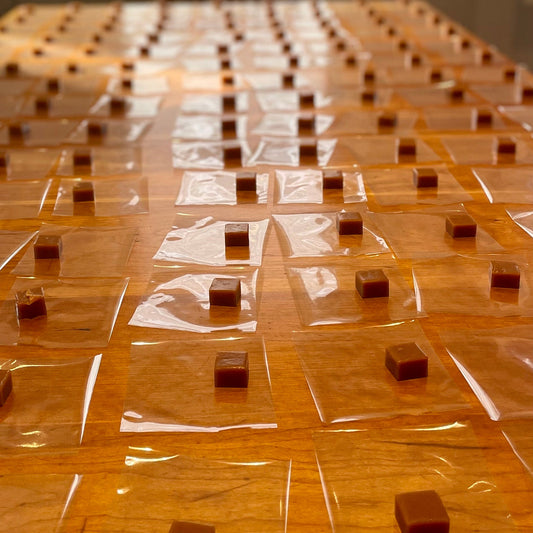 Small square caramels on clear cellophane square sheets on a wooden table, all in a row.