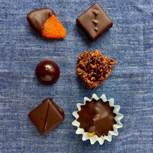 Overhead view of 6 items on denim blue material background: chocolate dipped apricot, chocolate dome, rocher, hazelnut + candied orange cup, 2 dark chocolate bonbons.